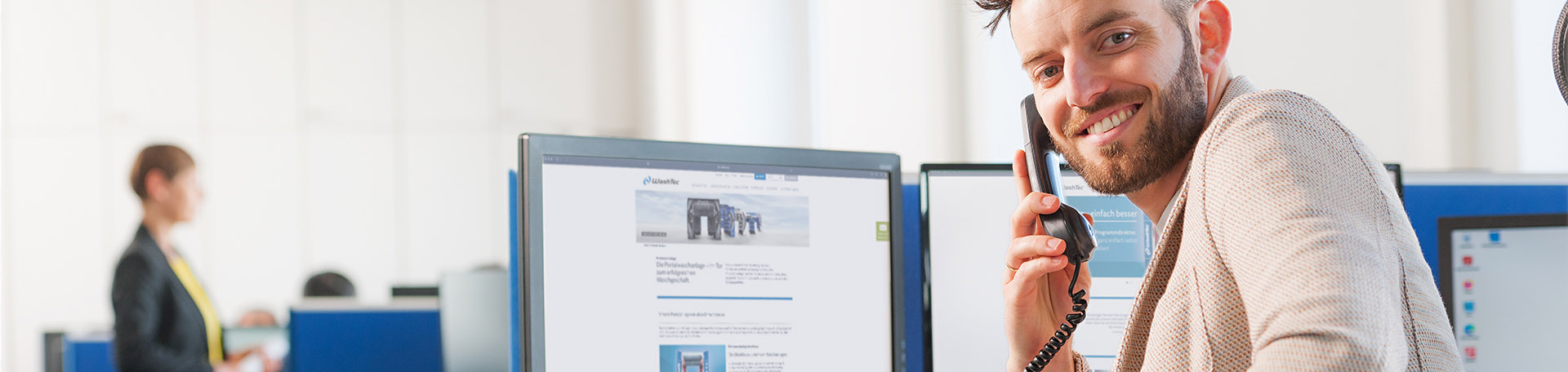 Eine Person sitzt mit Telefon am Ohr am Computer und lächelt in die Kamera.