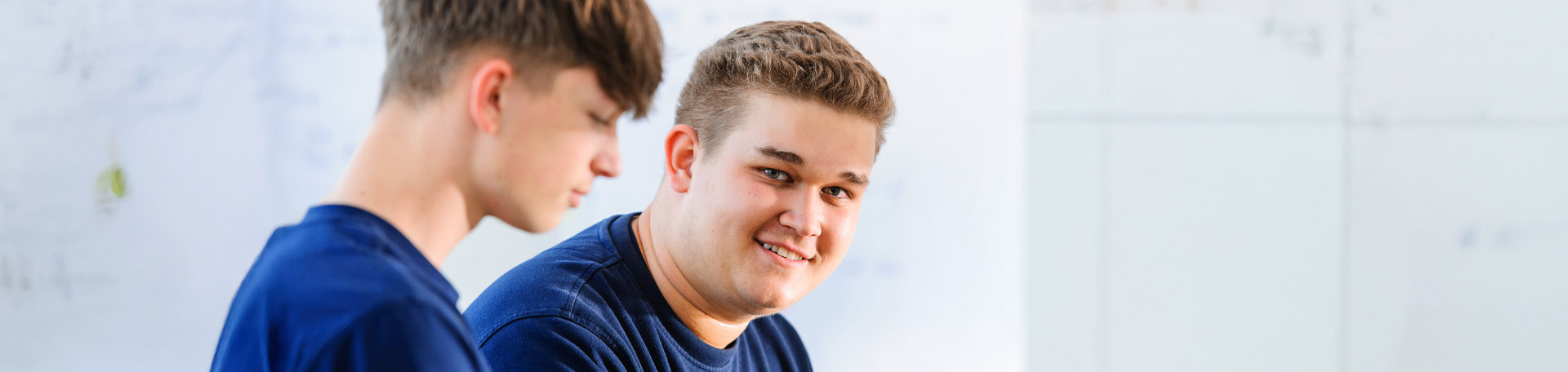 Zwei junge Personen sitzen nebeneinander seitlich zur Kamera. Die hintere Person blickt nach rechts und lächelt  in die Kamera.